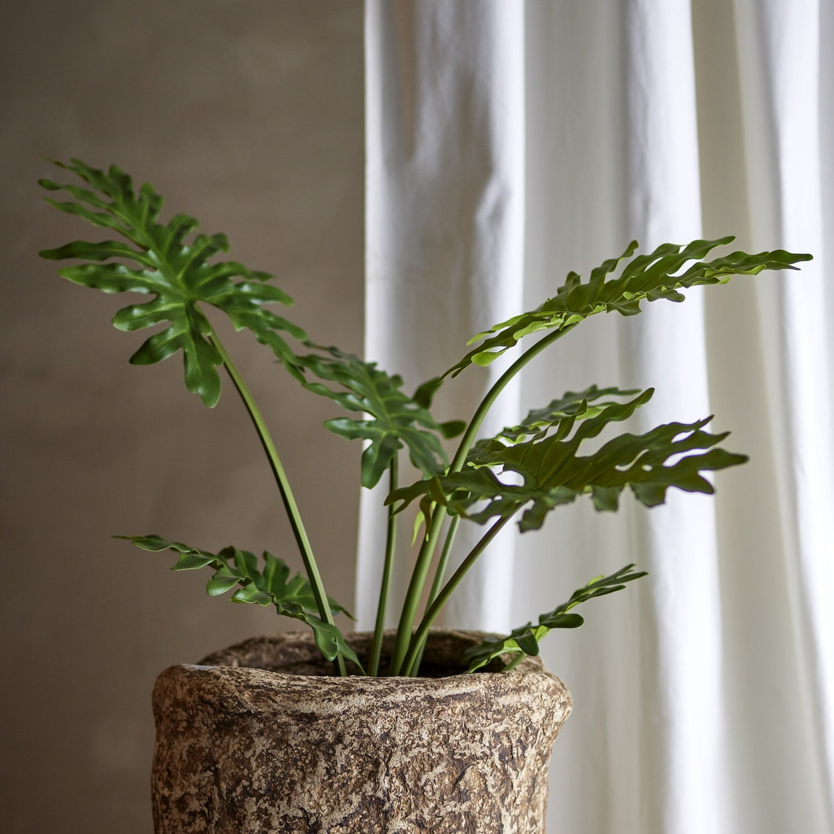 Philodendron Artificial Plant in Green, showing deeply cut leaves on upright stems in a textured stone-effect pot, set against a white curtain backdrop.