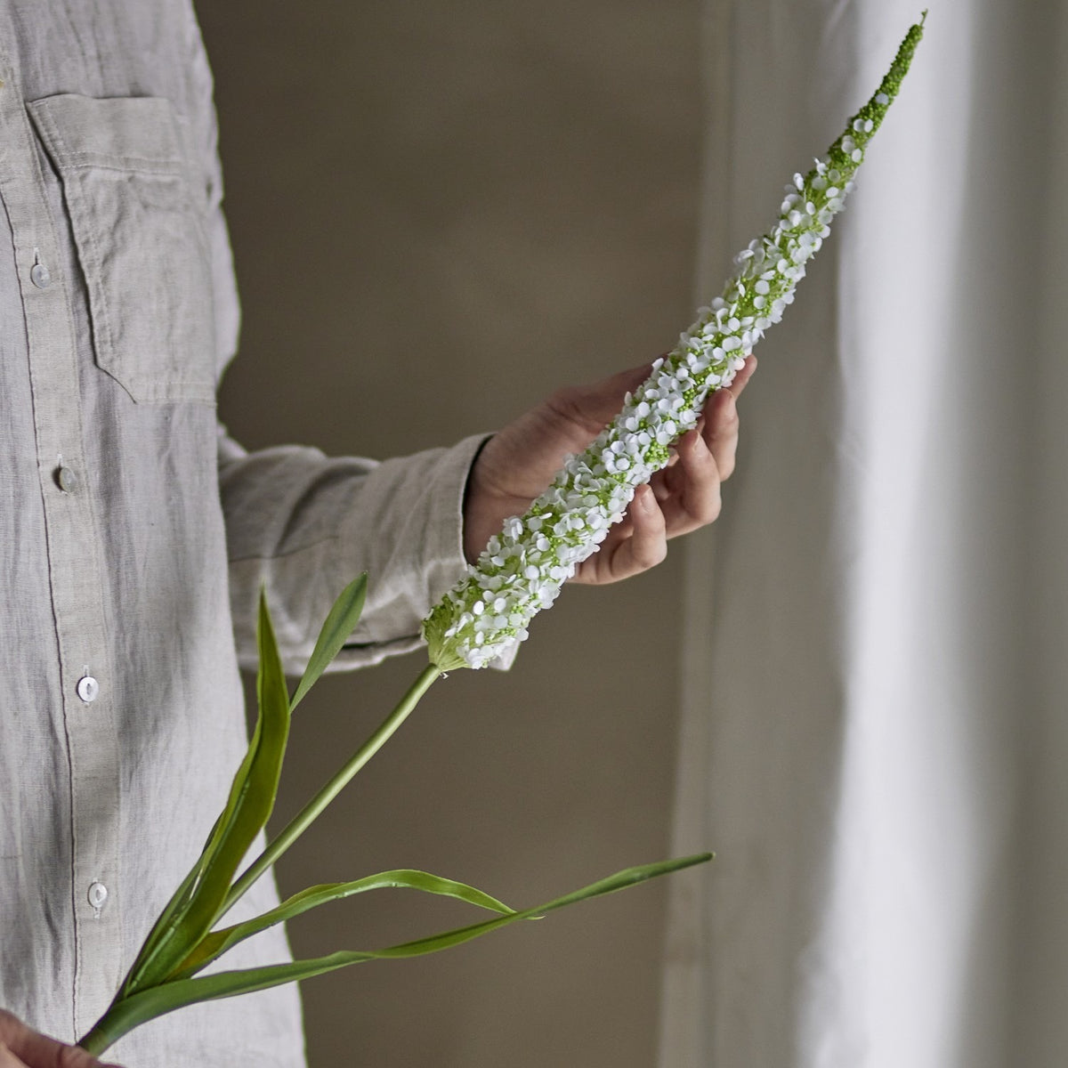 Foxtail Artificial Stem in White, held in hand showing the tapering spike of tiny white florets with green leaves below, set against a neutral indoor backdrop.