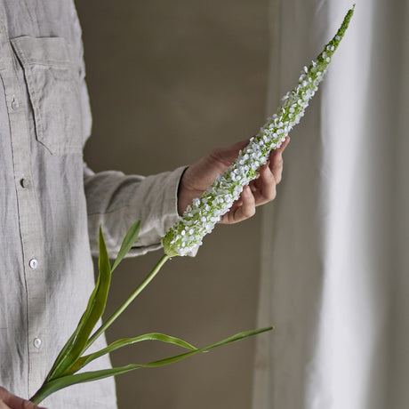 Foxtail Artificial Stem in White, held in hand showing the tapering spike of tiny white florets with green leaves below, set against a neutral indoor backdrop.