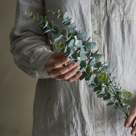 Eucalyptus Artificial Stem 76cm in green, held by hand against a neutral shirt, showing slender stem with small rounded leaves along its length.