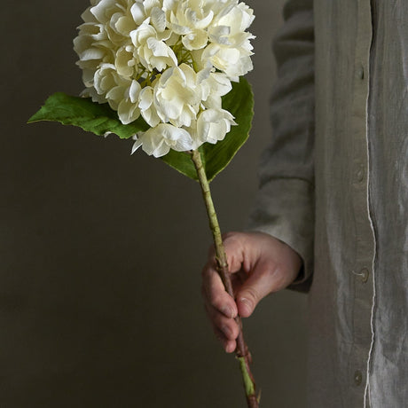 Hydrangea Artificial Stem in Cream held by hand, showing a rounded cream floret head, broad green leaves, and a knotted stem against a plain dark background.