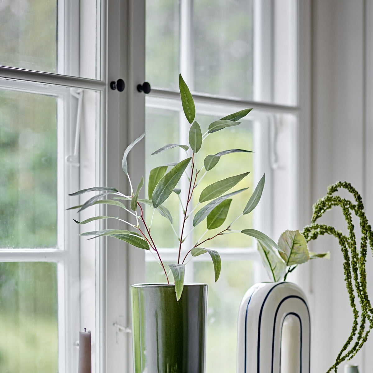 Eucalyptus Artificial Stem 85x18cm in Green displayed in a glossy green vase on a windowsill, its branching stalk and slim pointed leaves set against white-framed windows.