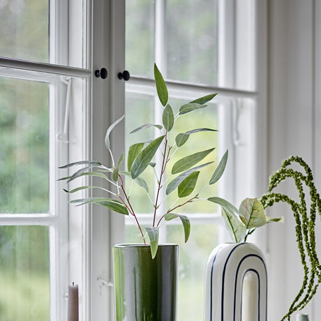 Eucalyptus Artificial Stem 85x18cm in Green displayed in a glossy green vase on a windowsill, its branching stalk and slim pointed leaves set against white-framed windows.