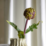 Protea Artificial Stem 54cm in purple, showing a layered bract flower head on a purple stem with two deeply cut leaves, displayed in a ridged cream vase against a curtained background.
