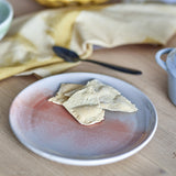 Stoneware side plate from the Paula Breakfast Set in orange reactive glaze, shown with crackers on a wooden table beside a spoon and linen napkin.