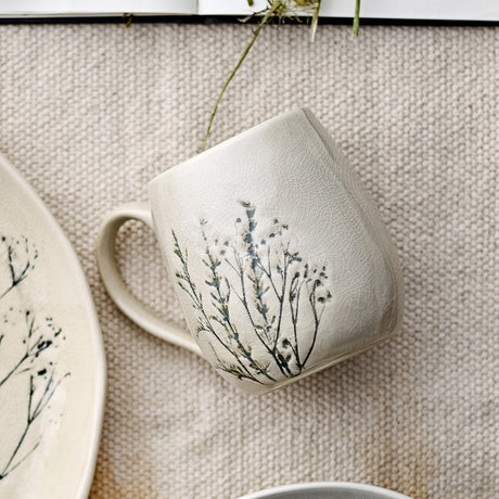 Cream stoneware mug from the Bea Breakfast Set of 12, with a rounded handle and crackle glaze finish featuring a floral sprig motif, shown alongside a matching plate on a woven neutral surface.