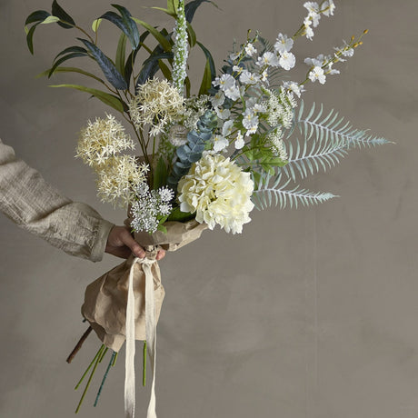 White Symphonie artificial bouquet held in hand, featuring a hydrangea-style flowerhead, lacy sprays, fern fronds and eucalyptus-style leaves, wrapped in paper with a ribbon tie.