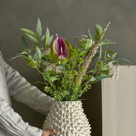 Person carrying a textured white vase holding the Blush Artificial Bouquet, featuring a pink heart-shaped bloom, green foliage, thistle heads and flowering sprigs against a muted grey wall.