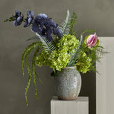 Poetry Artificial Bouquet in Green, featuring blue-toned orchid sprays, green hydrangea heads, a pink anthurium and fern fronds, arranged in a textured ceramic vase against a grey background.