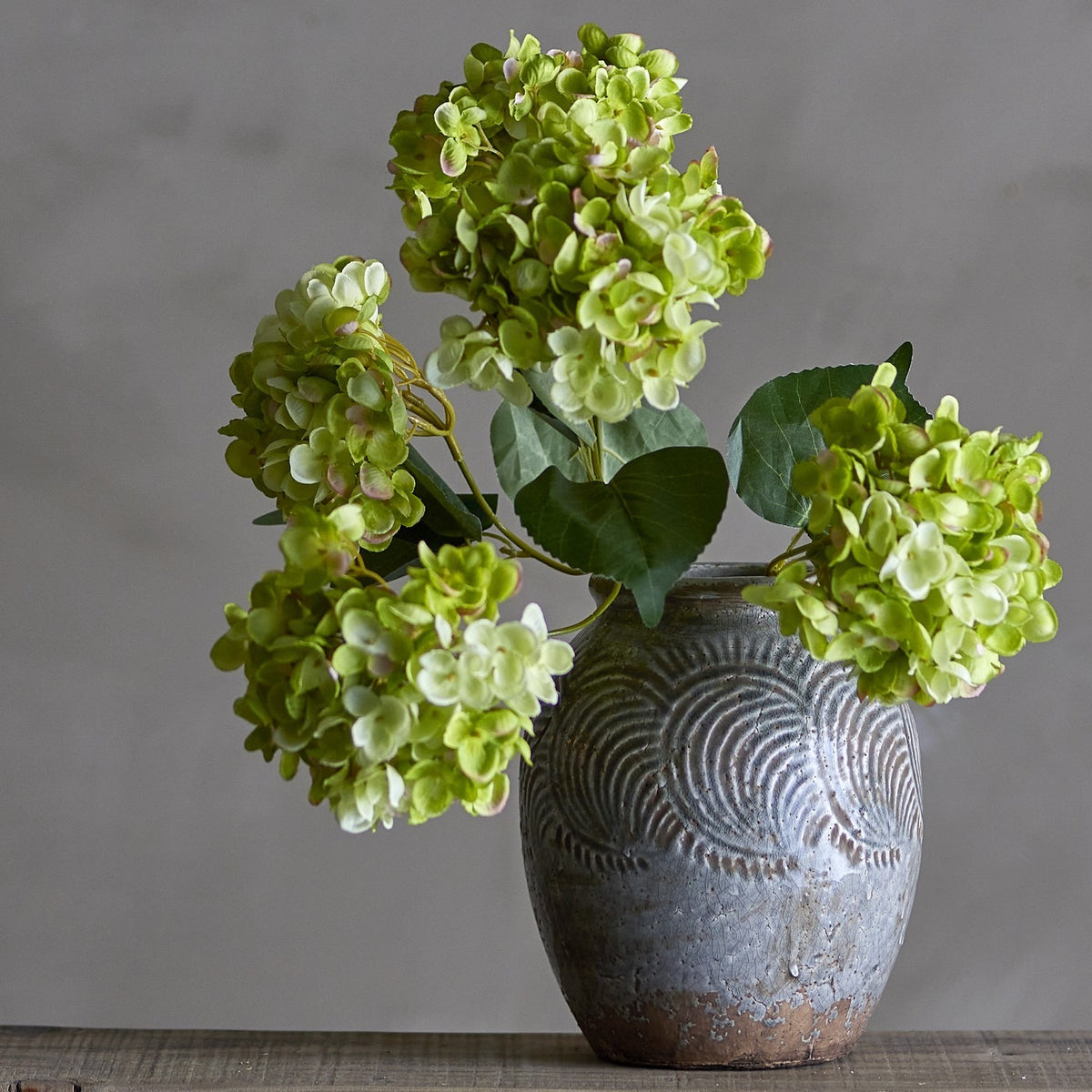 Hydrangea Artificial Stem 49cm in green, with multiple domed flowerheads and leafy stems arranged in a textured stoneware vase against a grey background.