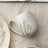 Bea Floral Stoneware Mug in cream with a crackled glaze and botanical branch design, shown with its loop handle beside a matching floral plate on a woven neutral surface.