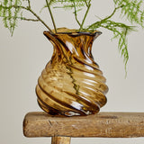Rylo Glass Vase 18.5cm in brown with spiralling ribs, a pinched neck and flared wavy rim, shown with greenery on a wooden stool against a plain background.