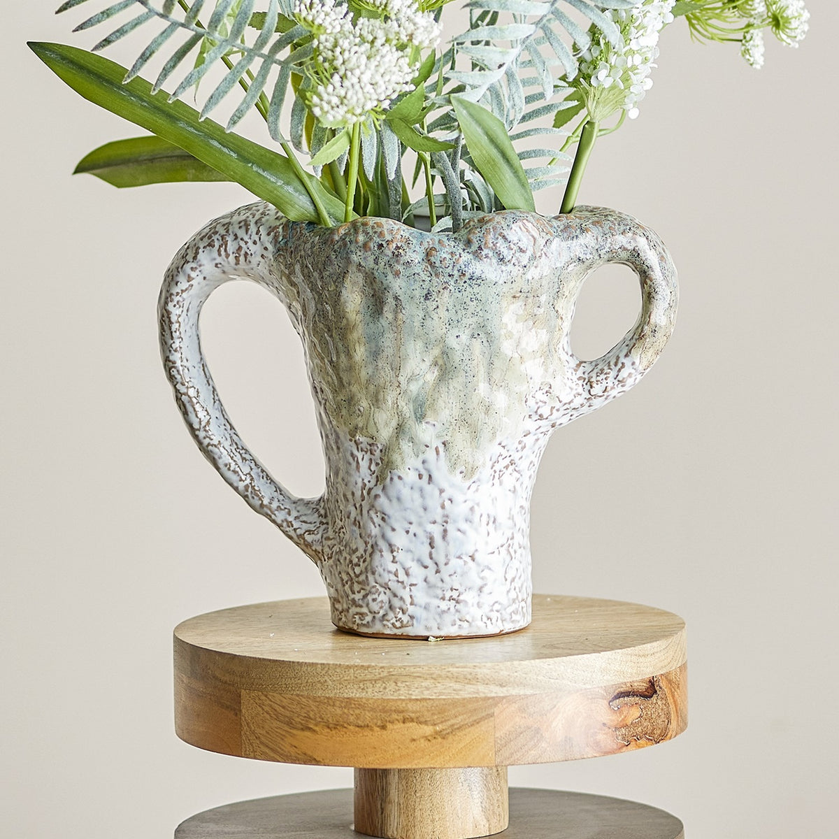 Ledger Speckled Stoneware Vase 21cm in White with wavy rim and two different-sized loop handles, holding greenery and white florals on a round wooden stand.