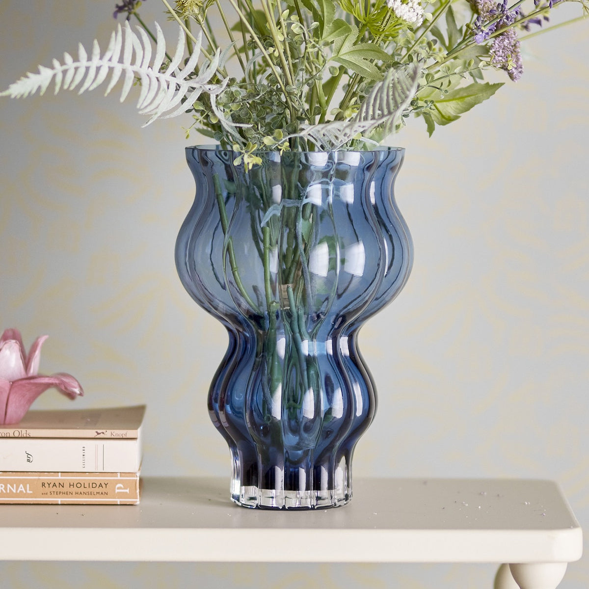 Zanna Glass Vase 26cm in blue with ribbed, wavy silhouette holds wildflowers on a pale shelf against a patterned wall, styled beside books and a decorative pink flower.