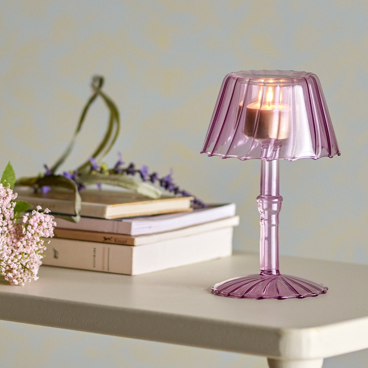 Cisco Glass Tealight Holder 18cm in pink, lamp-shaped with fluted shade and ribbed stem on a fluted foot, lit and displayed on a table beside books and flowers.