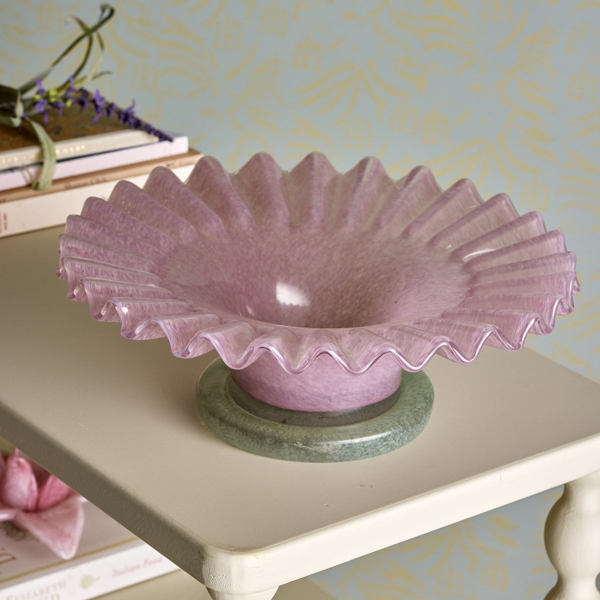 Garnet Glass Bowl 29cm in pink with a ruffled, ribbed rim, set on a green marble pedestal base atop a side table styled with books and florals.