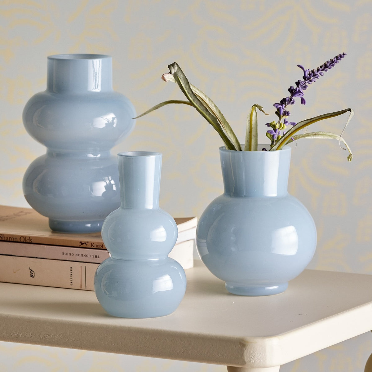 Kinsie Glass Vases Set of 3 in blue, each with stacked rounded bodies and varying neck heights, displayed on a white side table with books and a sprig of lavender.