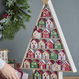 Calvin Christmas Tree Advent Calendar in MDF, triangular wooden frame with star top and 24 numbered houses in red, green and white, shown on a sideboard with festive greenery.