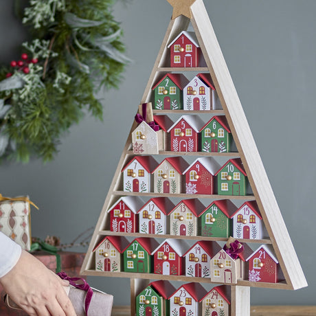 Calvin Christmas Tree Advent Calendar in MDF, triangular wooden frame with star top and 24 numbered houses in red, green and white, shown on a sideboard with festive greenery.