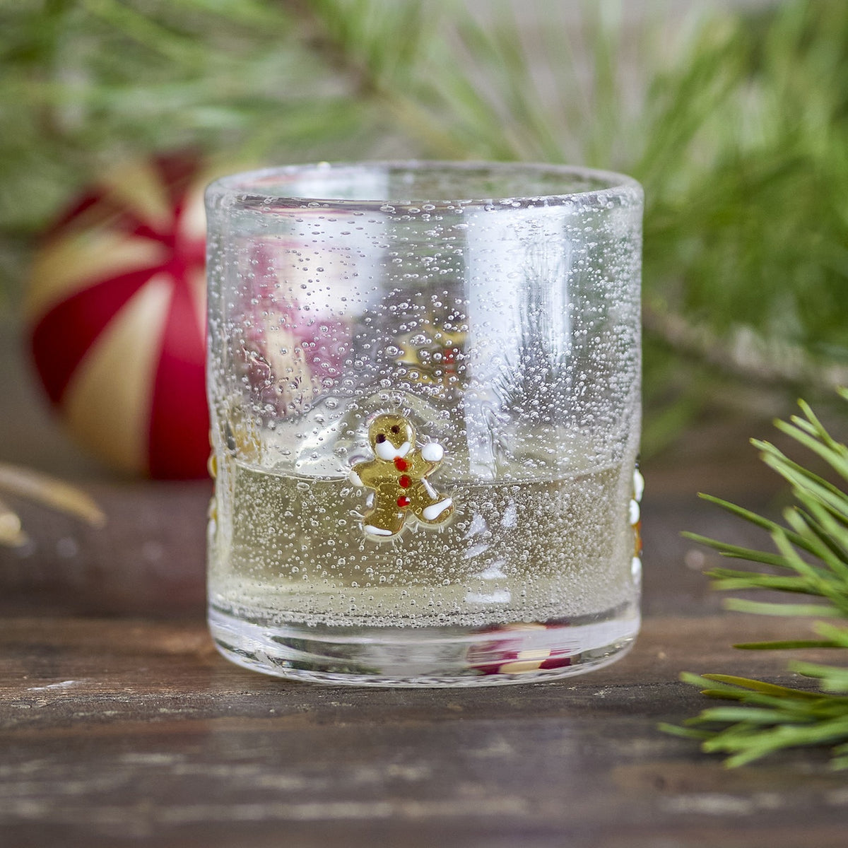Debra Gingerbread Christmas Tumbler in clear mouthblown glass with bubbled texture and a hand-painted gingerbread figure, shown with sparkling water and festive greenery in the background.