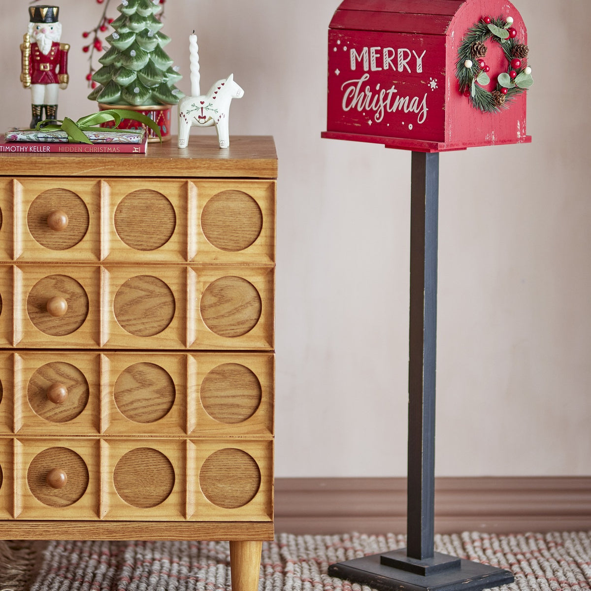 Carlo Christmas Decoration in red: freestanding postbox-style MDF ornament with curved slatted roof, "Merry Christmas" script, and a small pine wreath, shown beside a wooden drawer unit.