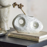 Caia Pebble Stoneware Vase 9.5cm in White, a textured pebble-shaped vase with two round cutouts, displayed with a dried stem atop books on a dark table.