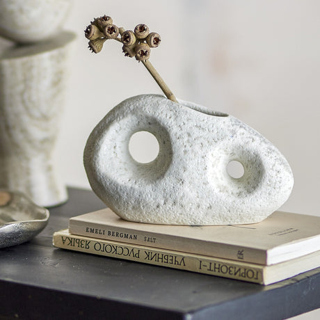 Caia Pebble Stoneware Vase 9.5cm in White, a textured pebble-shaped vase with two round cutouts, displayed with a dried stem atop books on a dark table.