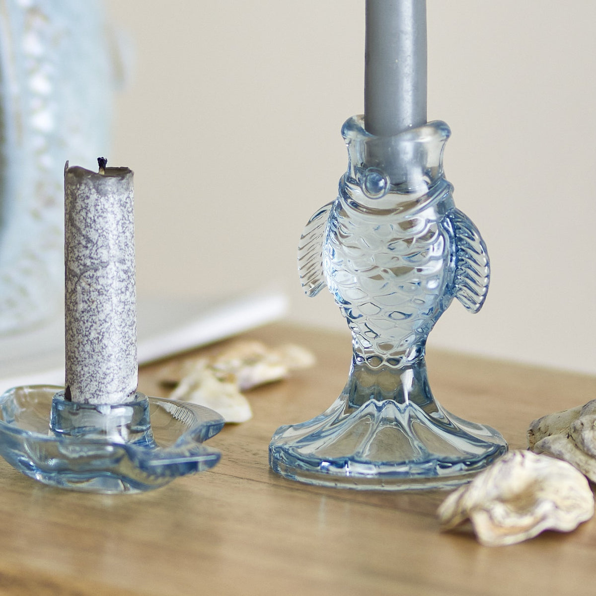 Linley Fish Glass Candle Holders in blue, one upright fish-shaped holder with a candle in its mouth and one flat fish dish holder, shown on a wooden surface with shells.