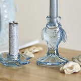 Linley Fish Glass Candle Holders in blue, one upright fish-shaped holder with a candle in its mouth and one flat fish dish holder, shown on a wooden surface with shells.