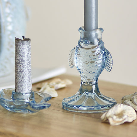 Linley Fish Glass Candle Holders in blue, one upright fish-shaped holder with a candle in its mouth and one flat fish dish holder, shown on a wooden surface with shells.