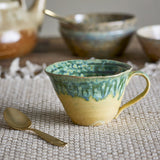 Milani Green Stoneware Cup 250ml, a wide, flared ceramic teacup with a mustard-yellow lower body and green reactive glaze dripping from the rim, shown with a gold spoon and bowls in the background.