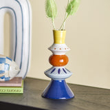 Kairi Stoneware Vase 21cm in multicoloured ceramic with stacked yellow, white, orange, blue and navy forms, holding dried foliage on a dark shelf beside books and a fish ornament.