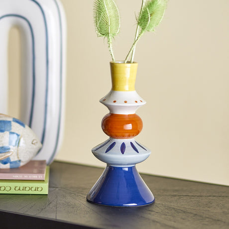 Kairi Stoneware Vase 21cm in multicoloured ceramic with stacked yellow, white, orange, blue and navy forms, holding dried foliage on a dark shelf beside books and a fish ornament.