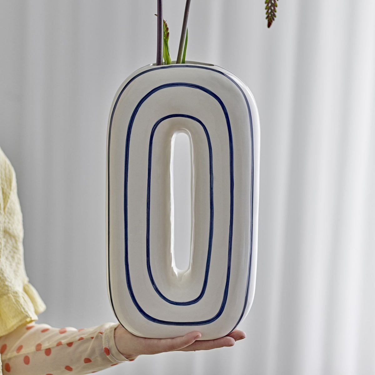 Mae Striped Ceramic Vase 32cm in White, an oval loop-shaped design with blue outlined rings, held in hand against a light curtain backdrop.