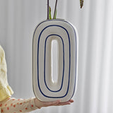 Mae Striped Ceramic Vase 32cm in White, an oval loop-shaped design with blue outlined rings, held in hand against a light curtain backdrop.