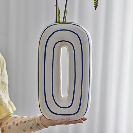 Mae Striped Ceramic Vase 32cm in White, an oval loop-shaped design with blue outlined rings, held in hand against a light curtain backdrop.