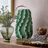 Natika Wavy Terracotta Vase in green, 37cm tall with ruffled wavy ridges, shown on a sideboard beside a glass vase and decorative accents in a living room setting.