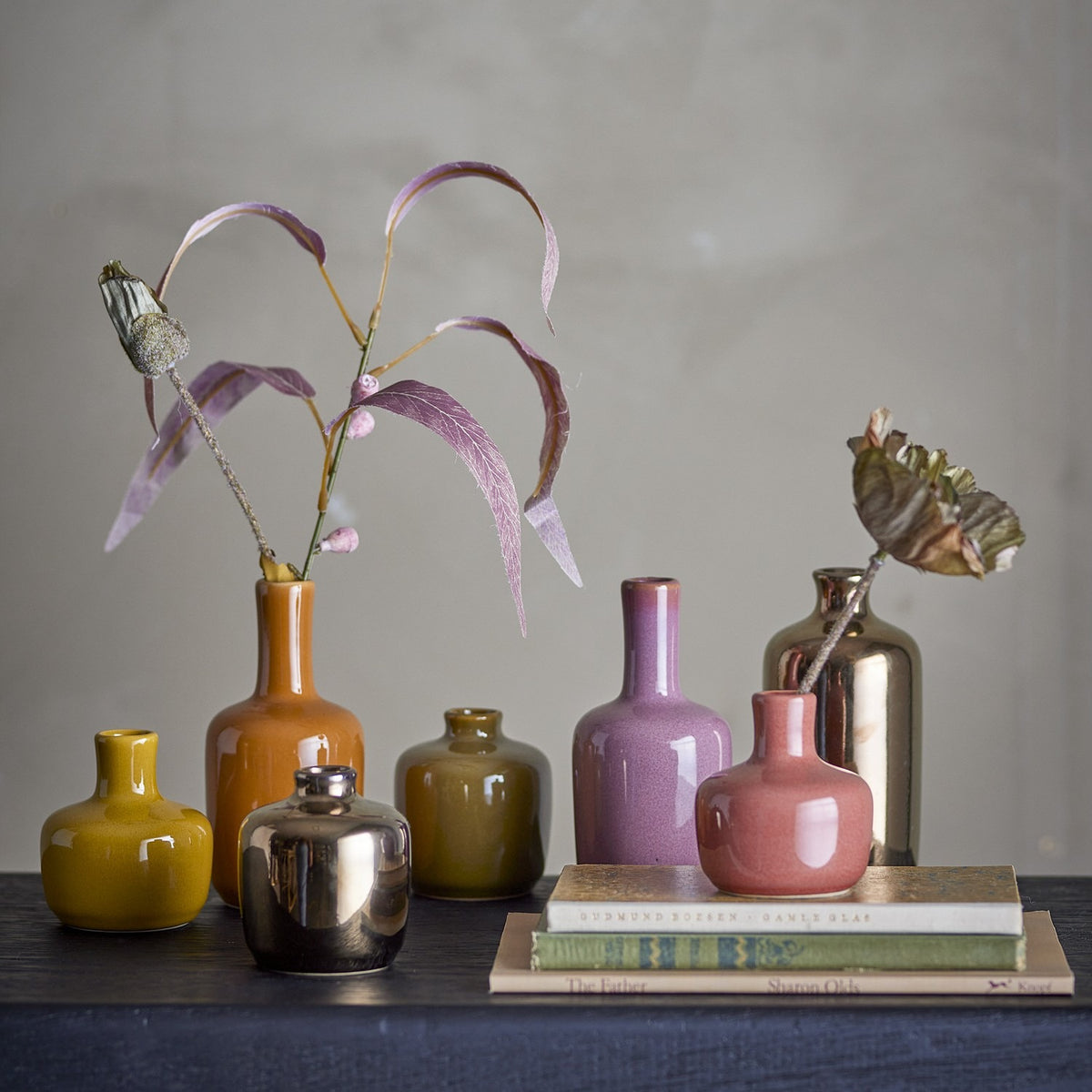 Maien Plain Stoneware Vases 14cm Set of 7 in mustard, orange, olive, pink, mauve and metallic finishes, holding dried leaves on a dark shelf with books.