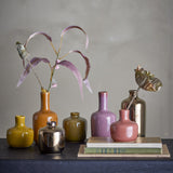 Maien Plain Stoneware Vases 14cm Set of 7 in mustard, orange, olive, pink, mauve and metallic finishes, holding dried leaves on a dark shelf with books.