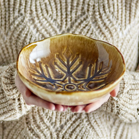 Acco Snowflake Stoneware Christmas Bowl 20cm in brown, glossy glaze with raised snowflake pattern inside, wavy rim, held in two hands against a knitted jumper.