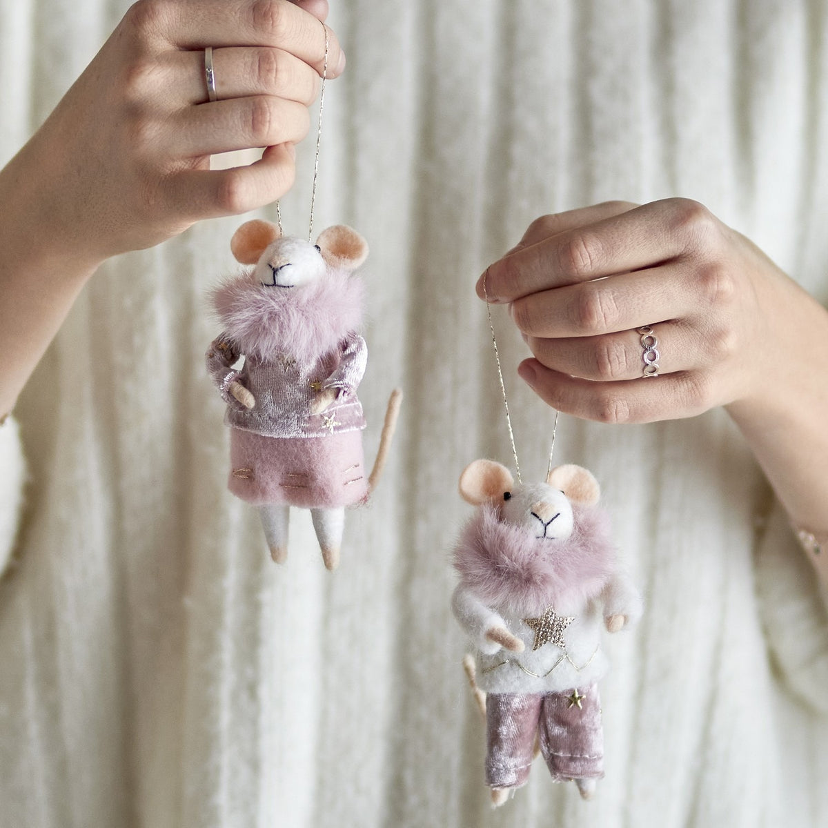 Hands holding two Peo Star felted wool mouse Christmas decorations in pink, one in a star-print jumper and trousers, the other in a top and skirt with fluffy collars, against a cream knit background.