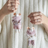 Hands holding two Peo Star felted wool mouse Christmas decorations in pink, one in a star-print jumper and trousers, the other in a top and skirt with fluffy collars, against a cream knit background.