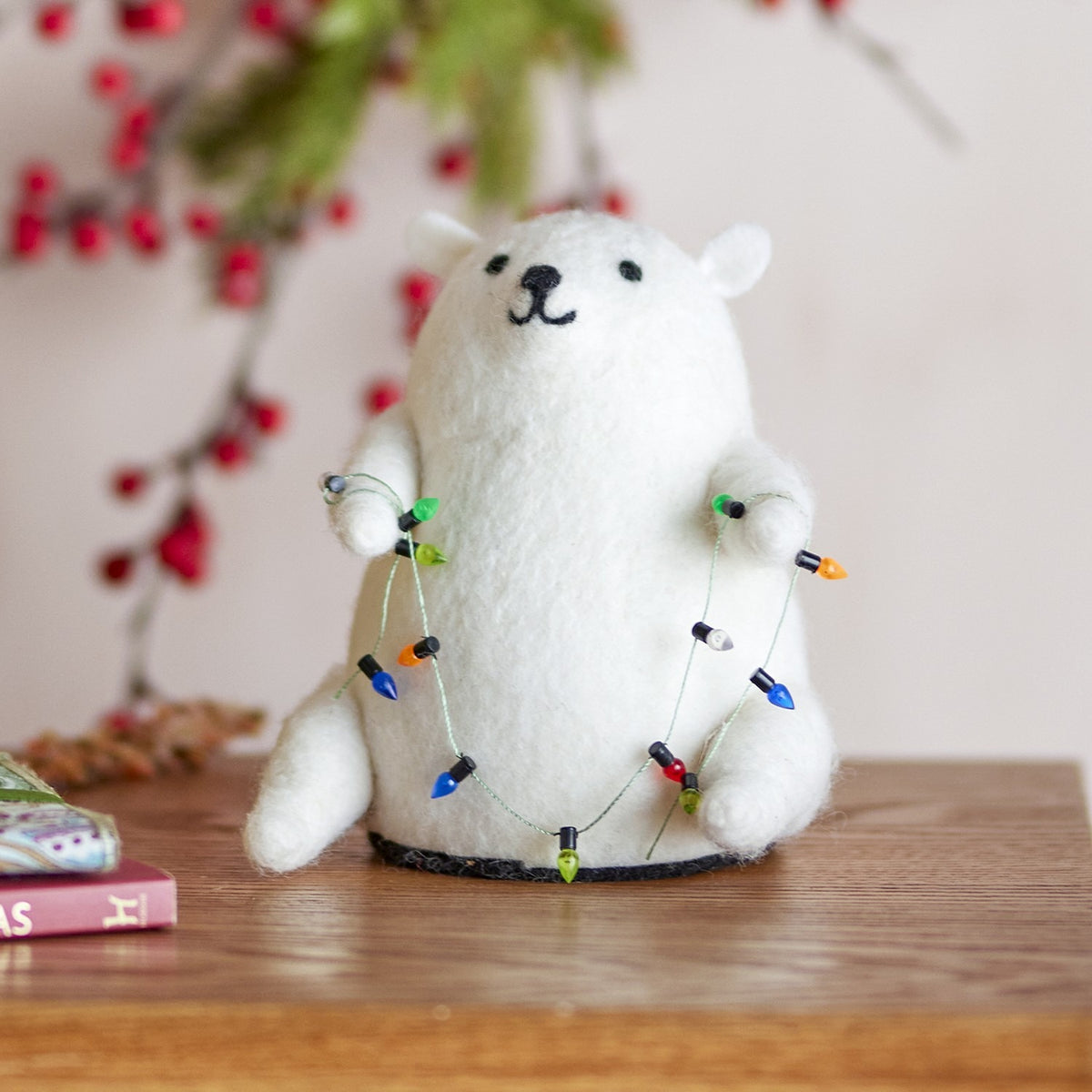 Wally Wool Christmas Decoration in white, a felted bear figure holding a string of colourful bulb lights, displayed on a wooden surface with festive berry foliage behind.