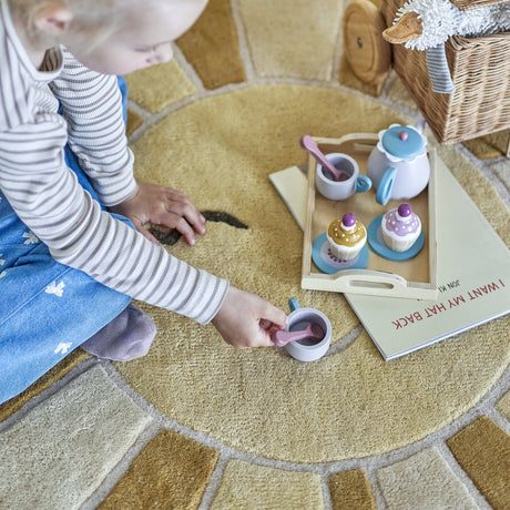 Child kneeling on the Sun Round Wool Rug 110cm in yellow, its sun-face design visible, while playing with a wooden toy tea set and cupcakes.