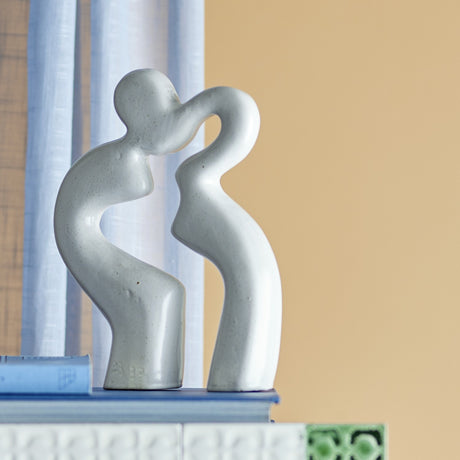 Sainab Stoneware Ornament in white, an abstract sculpture of two curving figures leaning together, shown close-up against a soft blue and tan backdrop.