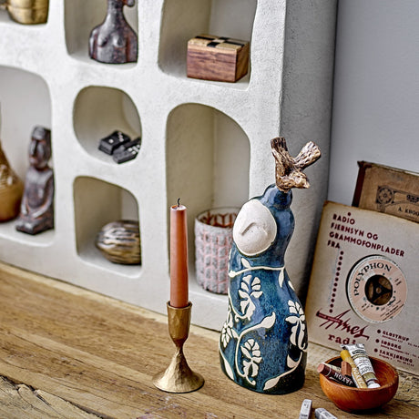 Layhan Stoneware Ornament in blue, a ceramic figurine with floral leaf patterns and a bird perched on its head, displayed on a wooden shelf beside a candle and books.