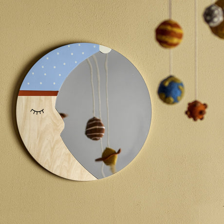 Moony Round Wooden Wall Mirror 40cm with a sleepy moon face, closed lashed eye and dotted nightcap, plywood crescent and mirrored glass, hung on a wall near a hanging felt planet mobile.