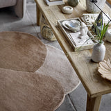 Denton Wool Rug in brown with overlapping pebble-shaped curves, shown beside a wooden coffee table styled with decor in a living room setting.
