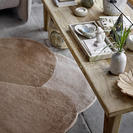 Denton Wool Rug in brown with overlapping pebble-shaped curves, shown beside a wooden coffee table styled with decor in a living room setting.