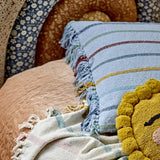Frey Striped Cotton Cushion in blue, woven with multicoloured stripes and fringed edges, shown on a bed with a plaid throw and a yellow pom-pom cushion.